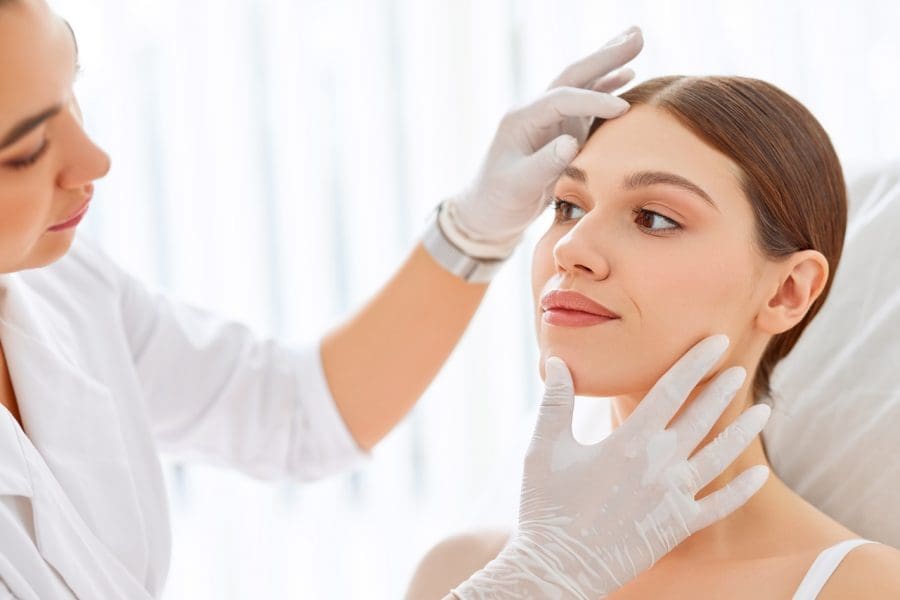 Crop cosmetologist in white gloves examining face skin of young female client before beauty procedure in salon